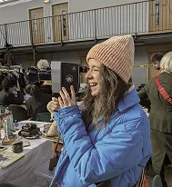 Maria outdoors at a flea market, laughing while holding a camera